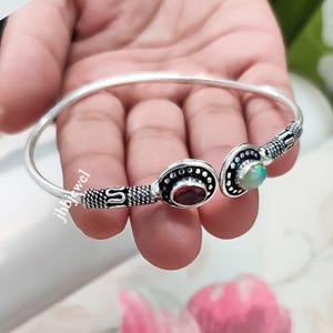May include: Silver-toned cuff bracelet featuring two oval settings. One setting holds a red gemstone, the other a light green gemstone. The bracelet has intricate detailing and is open at the wrist.