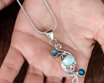 May include: A silver pendant necklace featuring a swirling design with a large, oval, iridescent stone and two smaller, round, blue stones. The necklace is held in a hand, showcasing the intricate details of the jewelry.
