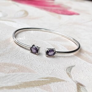 May include: A silver open cuff bracelet with two oval, deep purple gemstones. The bracelet has a simple, elegant design with the gemstones set at each end. The jewellery is displayed on a textured, off-white surface.