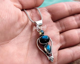May include: A silver pendant necklace featuring three iridescent blue gemstones. The pendant has a teardrop shape with a leaf design at the bottom. The text "jhjewel" is visible on the pendant. The necklace is held in a hand.