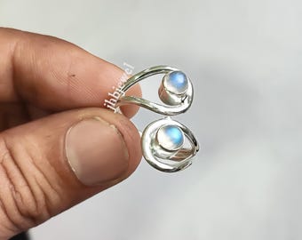 May include: A silver-colored ring with a unique open design, featuring two round, light blue moonstone cabochons. The ring's band curves around the stones, creating an elegant and modern look. The ring is held by a hand.