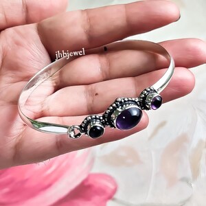 Op de afbeelding: Een zilveren armband met drie ovale paarse edelstenen en twee ronde zwarte edelstenen. De armband heeft een sierlijk ontwerp en de tekst "jhbjewel".