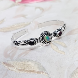 May include: A silver cuff bracelet featuring three gemstone accents. The center stone is an oval opal, flanked by a black onyx and a red garnet. The bracelet has an open design with decorative floral and dot patterns.