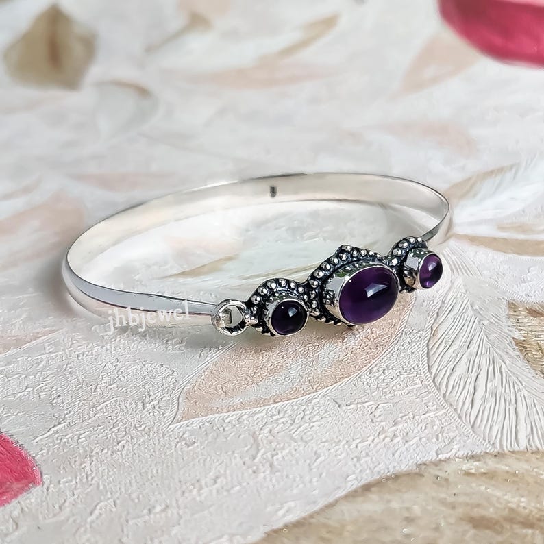 Op de afbeelding: Een zilveren armband met drie ovale en ronde paarse edelstenen. De armband heeft een decoratief ontwerp rond de edelstenen en is geplaatst tegen een getextureerde achtergrond. De merknaam "jhbjewel" staat op de armband.