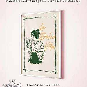 May include: A framed art print with a silver border. The artwork features a line drawing of a person holding a wine glass, with the words "La Dolce Vita" in gold script. The print has a cream background and a green decorative border.