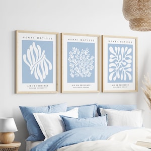 May include: Three framed art prints with light blue backgrounds and white abstract designs. Each print is labelled "HENRI MATISSE" and "AIX EN PROVENCE PAVILION DE VERSOME". The frames are a light wood colour.