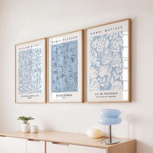 Może przedstawiać: Trzy oprawione grafiki z jasnoniebieskimi i białymi wzorami. Grafiki przedstawiają Williama Morrisa, Pablo Picasso i Henri Matisse. Ramki są w jasnym kolorze drewna. Grafiki są wyświetlane nad białą szafką.