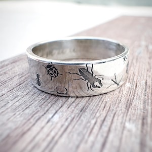 May include: A silver ring featuring etched insect illustrations, such as a ladybird, ant, and beetle. The ring has a broad band and a brushed metal finish. The insects are rendered in black, contrasting with the silver. The ring is presented on a wooden surface.