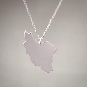 May include: A silver-colored necklace featuring a pendant shaped like the map of Iran. The pendant hangs from a delicate chain, creating a simple and elegant design. The necklace is displayed against a soft, neutral background.