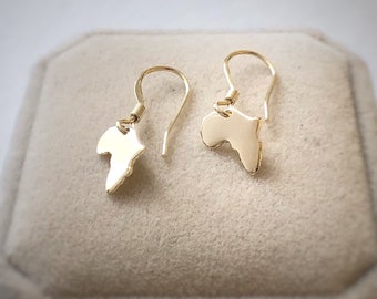 May include: A pair of gold-colored dangle earrings. Each earring features a small, solid silhouette of the African continent. The earrings are suspended from simple, curved ear wires. The earrings are displayed on a neutral-toned surface.