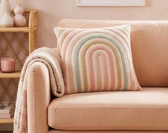 Pode incluir: Um sofá cor de pêssego com uma almofada com padrão de arco-íris e um cobertor estampado. Uma impressão emoldurada de um sol e uma lua está pendurada na parede. Uma prateleira tecida com itens decorativos está à esquerda, e um tapete de juta está no chão.