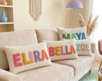 Pode incluir: Três almofadas retangulares de cor creme com superfícies texturizadas. Cada almofada apresenta letras de tecido grandes, coloridas e em relevo que soletram os nomes ELIRA, BELLA e ZOE, com uma almofada adicional atrás exibindo o nome MAYA.