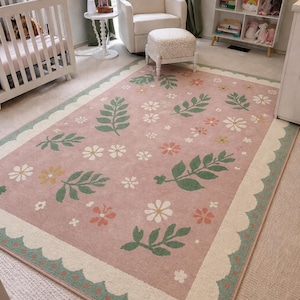 May include: A rectangular area rug featuring a dusty pink background with a repeating pattern of green leaves and small white, yellow, and coral flowers. The rug has a cream-colored scalloped border with small green dots along the edge.
