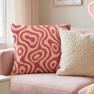 Op de afbeelding: Een vierkant sierkussen met een getextureerd punch-needle ontwerp met concentrische, golvende patronen in magenta en crème. Daarnaast ligt een effen, crèmekleurig kussen met een hoogpolige sherpa-textuur op een zachtroze bank.