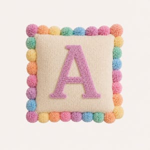 Pode incluir: Uma almofada quadrada de cor creme com uma letra A texturizada e acolchoada em tom lavanda no centro. As bordas da almofada s&atilde;o decoradas com uma borda de pequenos pompons multicoloridos em tons de rosa, amarelo, azul e laranja.