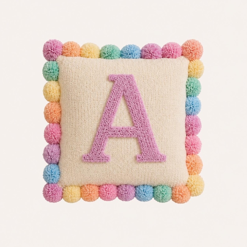 Pode incluir: Uma almofada quadrada de cor creme com uma letra A texturizada e acolchoada em tom lavanda no centro. As bordas da almofada s&atilde;o decoradas com uma borda de pequenos pompons multicoloridos em tons de rosa, amarelo, azul e laranja.