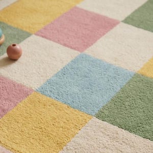 Soft pastel checkerboard nursery rug with scalloped border in pink, sage green, cream, yellow, and baby blue, styled in a cozy rainbow-themed kids room with plush tufted texture.