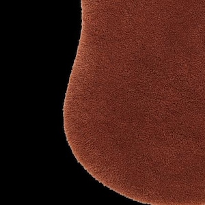 May include: Close-up of a textured, rust-colored, rounded object against a black background. The object has a soft, plush appearance, suggesting a rug or textile. The edges are slightly curved.