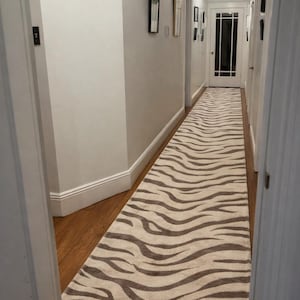 Könnte beinhalten: Ein Flurläufer-Teppich mit Zebra-Streifenmuster in Beige- und Brauntönen. Der rechteckige Teppich bedeckt die Länge eines Flurs mit Holzboden. Die Wände sind hell gestrichen, und es gibt gerahmte Bilder.