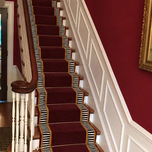 Peut inclure: Un escalier doté d'un chemin de tapis rouge foncé avec une bordure rayée noire et blanche et des barres d'escalier en métal doré. L'escalier présente des balustres en bois blanc, une rampe en bois foncé et des lambris blancs sur des murs rouges.