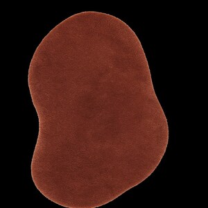 May include: A solid, irregularly shaped rug in a warm, reddish-brown hue. The rug has a soft, textured surface and is set against a stark black background. The design is abstract and modern, suitable for various interior styles.