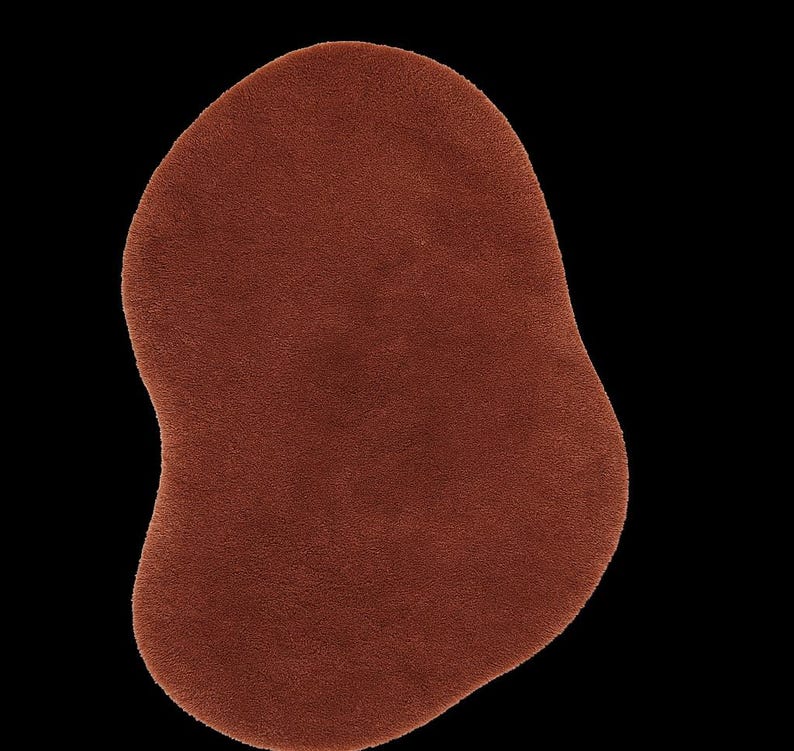 May include: A solid, irregularly shaped rug in a warm, reddish-brown hue. The rug has a soft, textured surface and is set against a stark black background. The design is abstract and modern, suitable for various interior styles.
