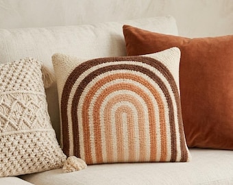 Pode incluir: Uma almofada quadrada com um design texturizado de punch needle representando um arco-íris em tons de creme, laranja claro e castanho escuro. A almofada tem um tecido base de cor creme e inclui uma pequena borla decorativa no canto.