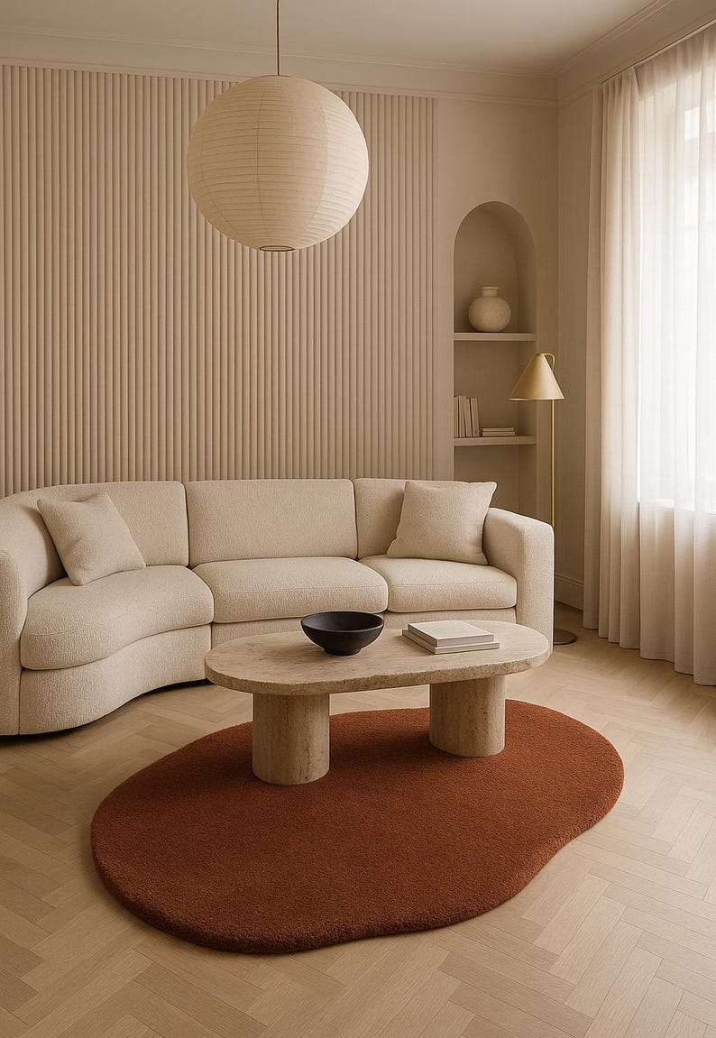 May include: A modern living room with a beige sectional sofa, a stone coffee table, and a rust-colored, organic-shaped rug. A paper lantern hangs from the ceiling, and a gold floor lamp stands near a window with sheer curtains. The walls are a neutral beige.