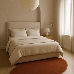 May include: A bedroom scene featuring a bed with beige bedding, pillows, and a headboard. A rust-colored, organic-shaped rug sits on the wooden floor. A paper lantern hangs from the ceiling, and a small lamp sits on a bedside table.