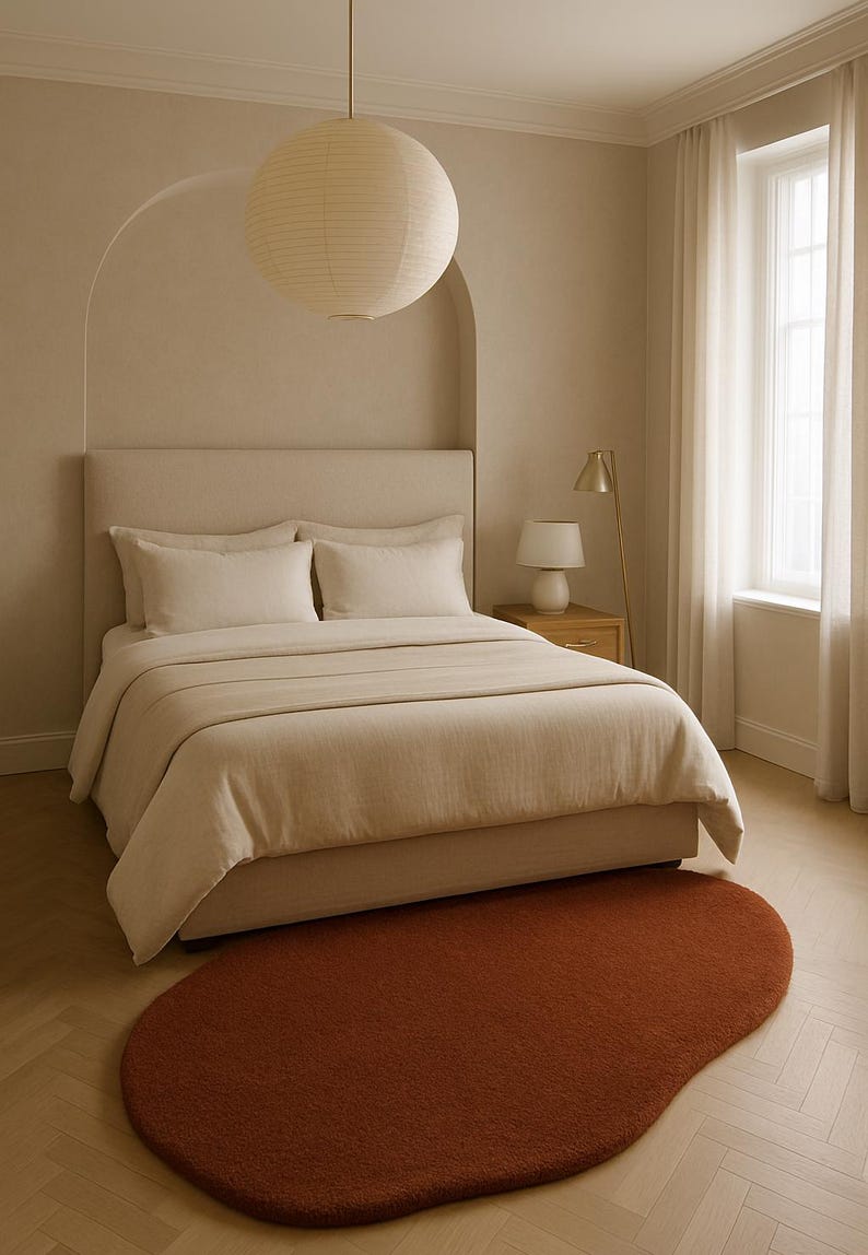May include: A bedroom scene featuring a bed with beige bedding, pillows, and a headboard. A rust-colored, organic-shaped rug sits on the wooden floor. A paper lantern hangs from the ceiling, and a small lamp sits on a bedside table.