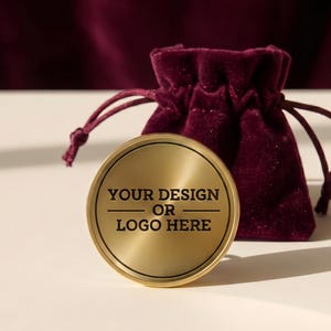 May include: A circular gold-toned metal coin with a brushed finish featuring the black text "YOUR DESIGN OR LOGO HERE" centered on the front, accompanied by a small, deep burgundy velvet drawstring pouch with matching cord ties.