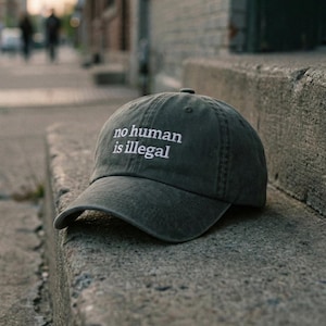 May include: A dark green baseball cap with the white text "no human is illegal" embroidered on the front. The cap is resting on a concrete step outdoors, with a blurred street scene in the background.