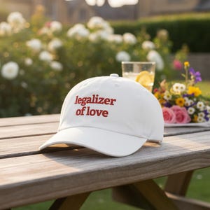 May include: A white baseball cap with the text "legalizer of love" in red script. The cap is sitting on a wooden picnic table outdoors, with flowers and a drink in the background.