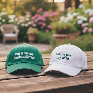 May include: Two baseball caps with text on a wooden surface. One is green with the text "Put it on my husband's tab" in white. The other is white with the text "Let me ask my wife" in green.