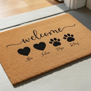 May include: A rectangular tan coir doormat featuring the black text welcome in a cursive script. Below the text are two black heart icons labeled Alex and Julia, followed by two black paw print icons labeled Max and Riley in black script.