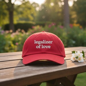 May include: A red baseball cap with the words "legalizer of love" embroidered in white. The cap is sitting on a wooden table with a sprig of white flowers. The background is a blurred view of a garden.
