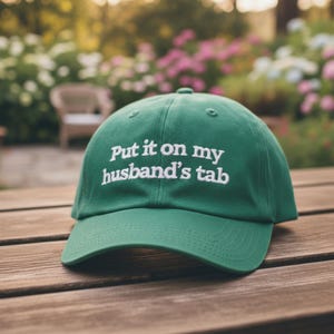 May include: A green baseball cap with the embroidered text "Put it on my husband's tab" in white. The cap is sitting on a wooden surface, with a blurred background of greenery and flowers.