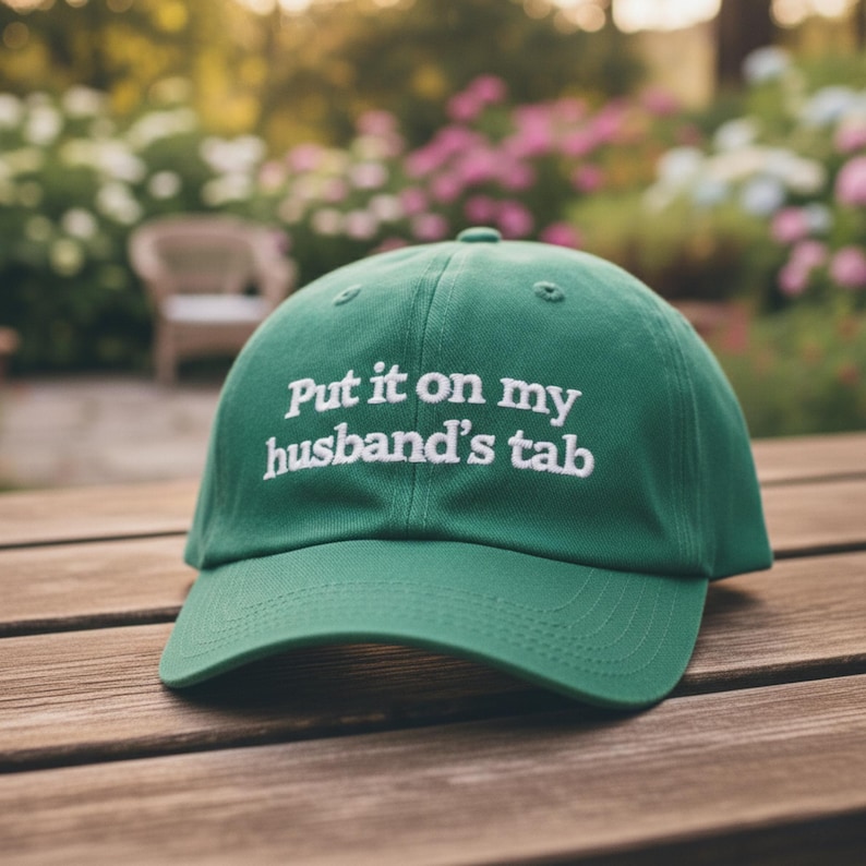 May include: A green baseball cap with the embroidered text "Put it on my husband's tab" in white. The cap is sitting on a wooden surface, with a blurred background of greenery and flowers.