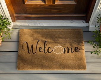 May include: A rectangular brown coir doormat with the word "Welcome" in a cursive font and a white outline pumpkin design. The mat is placed in front of a wooden door.
