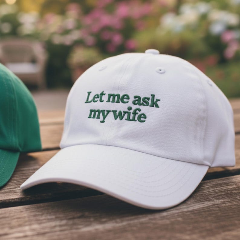 May include: White baseball cap with the embroidered text "Let me ask my wife" in green. The cap has a curved brim and a fabric strap closure. A green cap is partially visible on the left side of the image.