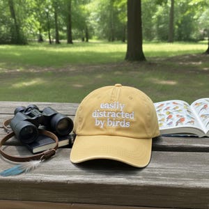 May include: Mustard yellow baseball cap with the embroidered text "easily distracted by birds." The cap is displayed with binoculars, a notebook, a bird book, and a blue feather on a wooden surface.
