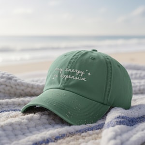 May include: A green baseball cap with the text "my energy is expensive" embroidered in white. The hat is resting on a striped towel on a beach. The background shows the ocean and sky.