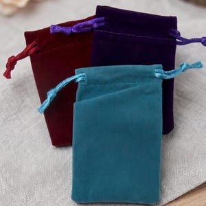 May include: Three small rectangular drawstring pouches made of soft velvet fabric. One pouch is deep teal with a matching light blue cord, one is dark burgundy with a red cord, and one is deep purple with a matching purple drawstring cord.