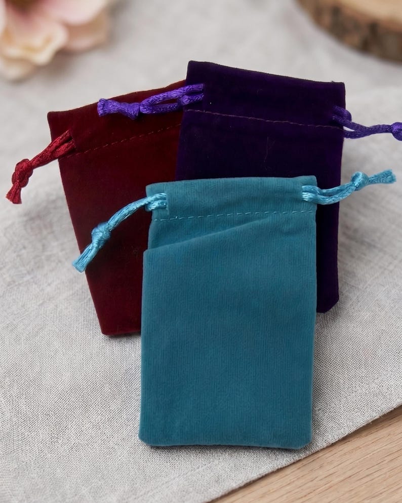 May include: Three small rectangular drawstring pouches made of soft velvet fabric. One pouch is deep teal with a matching light blue cord, one is dark burgundy with a red cord, and one is deep purple with a matching purple drawstring cord.