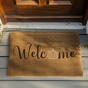 May include: A rectangular brown coir doormat featuring the word Welcome in a black cursive script. The letter o in the word is replaced by a white line drawing of a pumpkin. The mat has a coarse, fibrous texture and a natural tan color.