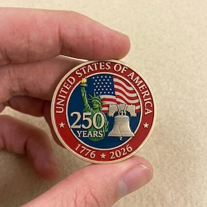 May include: A circular commemorative coin featuring the Statue of Liberty, the Liberty Bell, and an American flag. The text reads UNITED STATES OF AMERICA, 250 YEARS, and 1776-2026. The design uses red, white, blue, and gold metallic colors.
