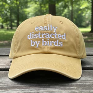May include: Mustard yellow baseball cap with the text "easily distracted by birds" embroidered in white. The cap has a curved brim and a classic six-panel design. The hat is made of a soft, durable fabric.