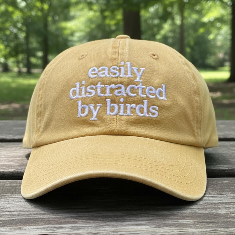 May include: Mustard yellow baseball cap with the text "easily distracted by birds" embroidered in white. The cap has a curved brim and a classic six-panel design. The hat is made of a soft, durable fabric.