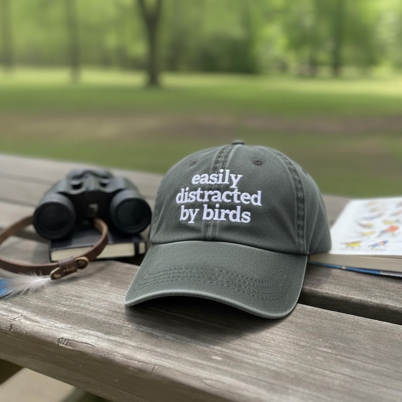 May include: A green baseball cap with the text "easily distracted by birds" in white. The cap is on a wooden picnic table next to binoculars, a notebook, and a bird guide. The background is a blurred outdoor scene.