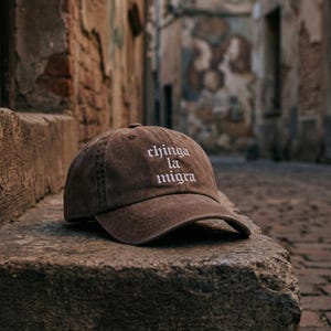 Può includere: Un cappellino da baseball marrone con la scritta ricamata "chinga la migra" in bianco. Il cappellino ha una visiera curva ed è realizzato in un materiale morbido e usurato. Il cappello è appoggiato su una superficie di pietra.